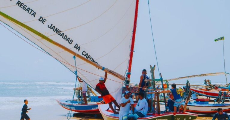 Jangadeiros reunidos na praia ao lado de jangada com vela personalizada, durante a Regata de Jangadas da Caponga no litoral do Ceará