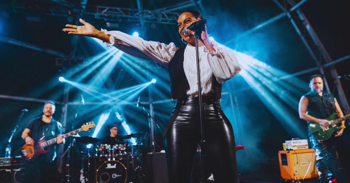 Cantora Alegria no palco durante tributo a Sade, com banda ao vivo e iluminação cênica colorida.