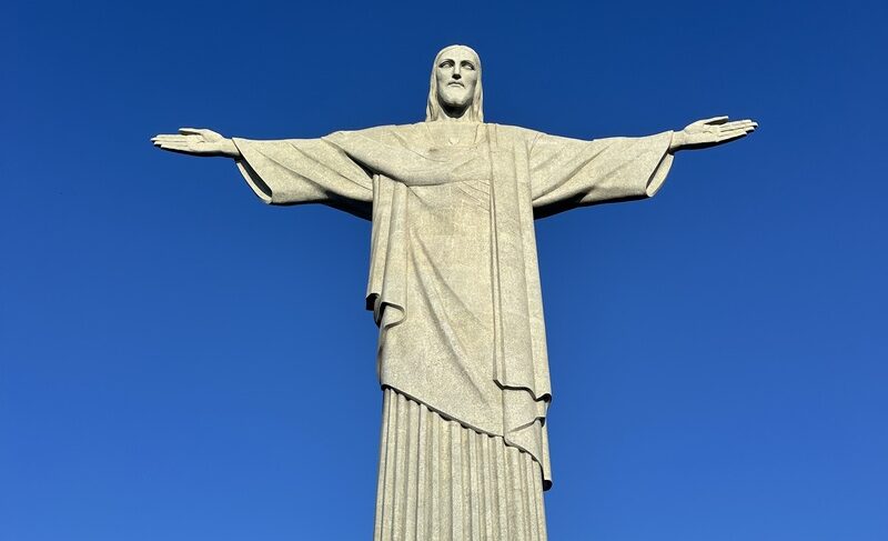 Vista do monumento do Cristo Redentor no topo do Morro do Corcovado, com a cidade do Rio de Janeiro e o Pão de Açúcar ao fundo.