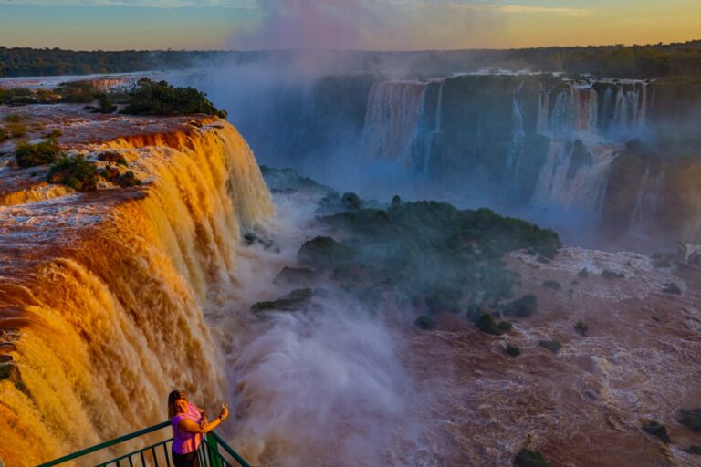 Vista panorâmica das quedas d'água das Cataratas do Iguaçu, com passarelas e turistas ao fundo, sob o sol do verão.