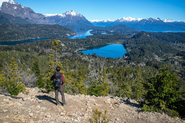 Paisagem de verão em Bariloche com lago cristalino, montanhas ao fundo e pessoas praticando caiaque.